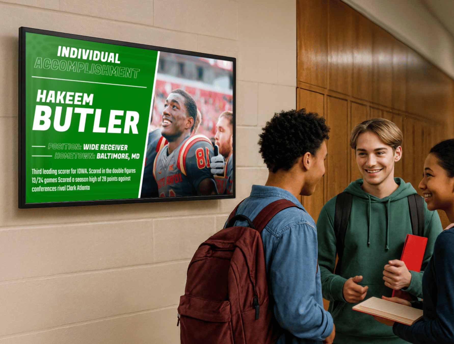 Students chatting in a school hallway with a digital hall of fame board mounted on the wall