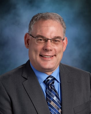 man with short professional grey hair wearing glasses smiling in grey suit with tie