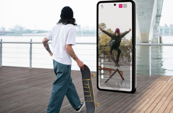 A man holding a skateboard watching skateboarding videos on a digital signage screen.