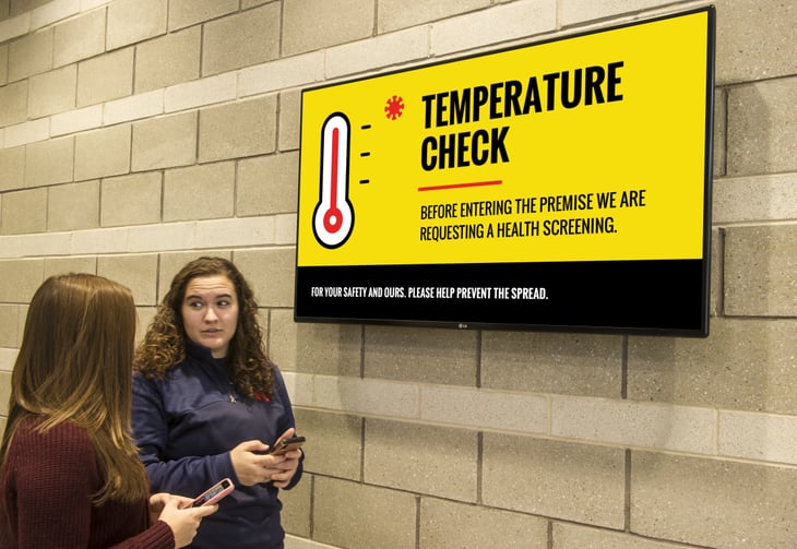 Two girls standing in front of a digital sign that says you need to get a temperature check