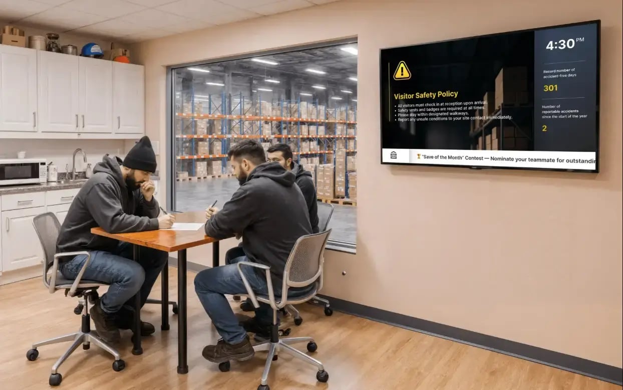 Warehouse workers inside an office of a warehouse with digital signage mounted on the wall