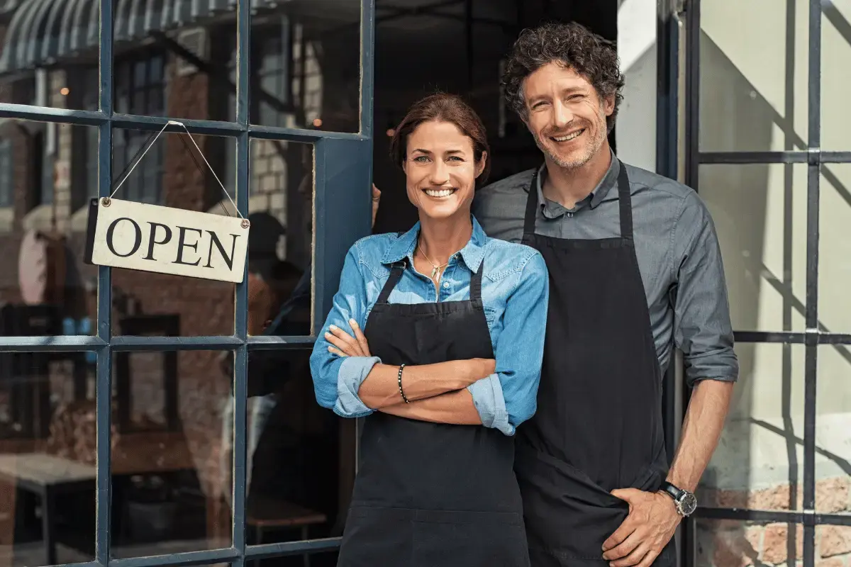 Two restaurant owners standing and smiling near their restaurant front door with an open sign hanging on the window