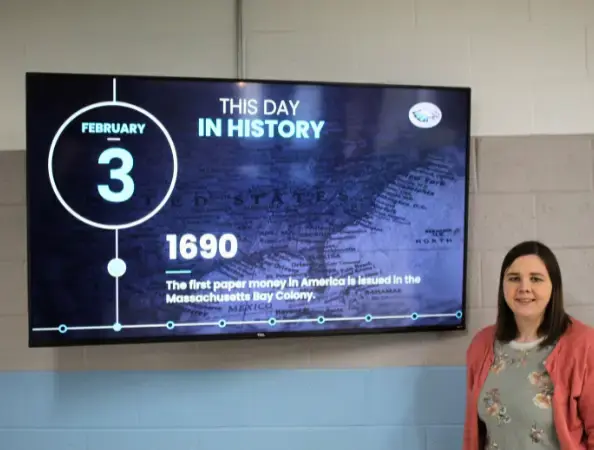 A female social media employee at a school district standing in front of a large TV displaying digital signage
