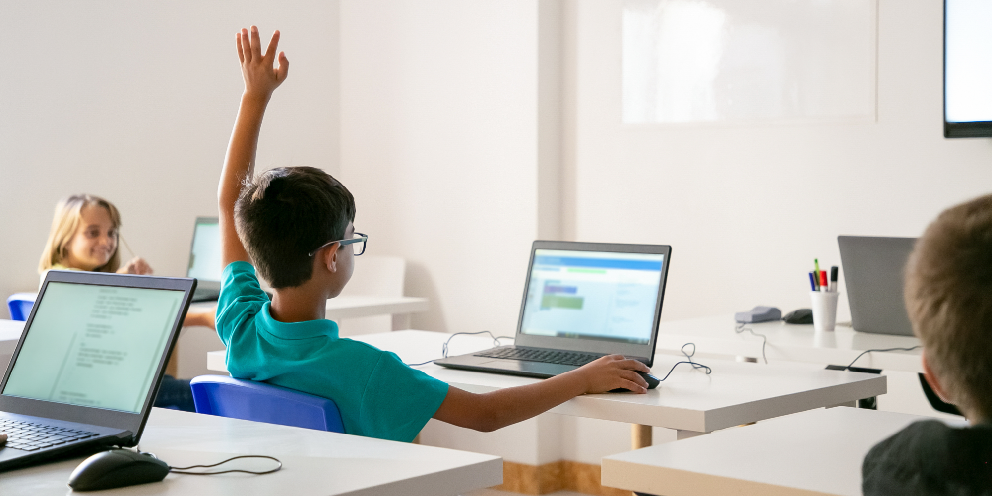 Student raising his hand with a laptop on desk.